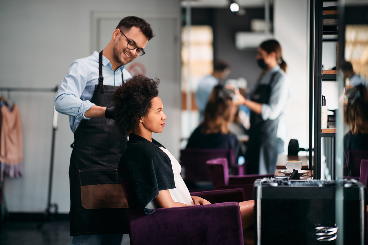 Happy male hairdresser styling hair of African American woman during appointment at the salon. Focus is on Sanepid control